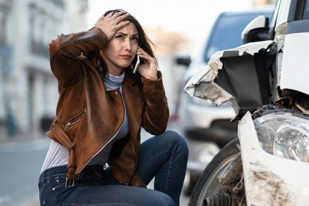 Distressed driver on the phone after a car accident, next to a damaged vehicle.
