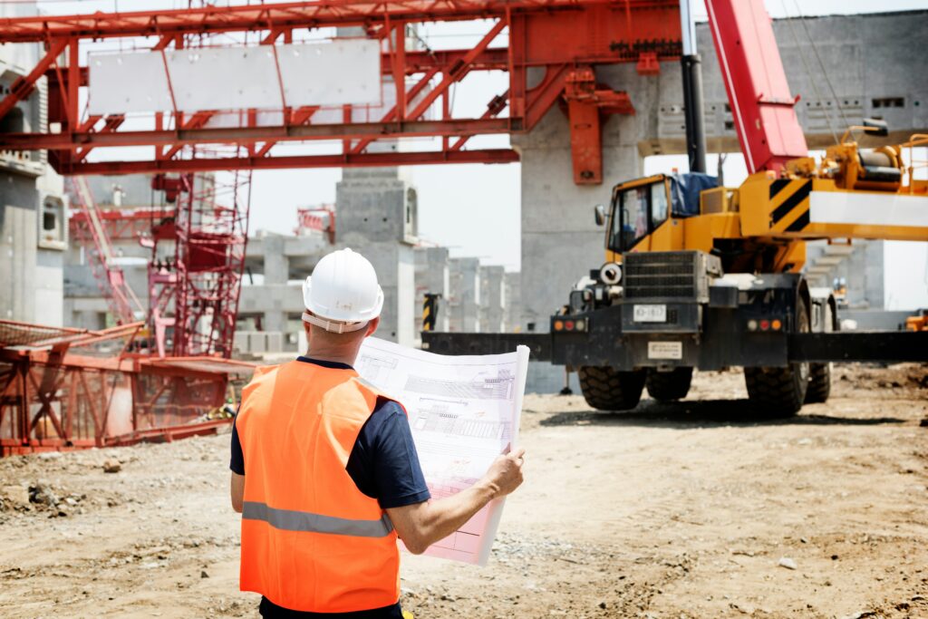 A construction supervisor in a hard hat and orange safety vest reviews blueprints at a building site with cranes and heavy machinery in the background.