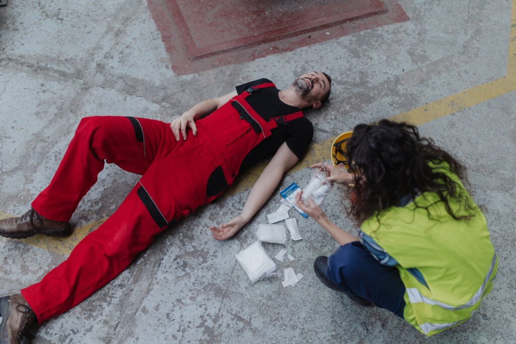 A worker in red coveralls lies injured on the ground while a colleague provides first aid at an industrial site, representing workplace accidents.