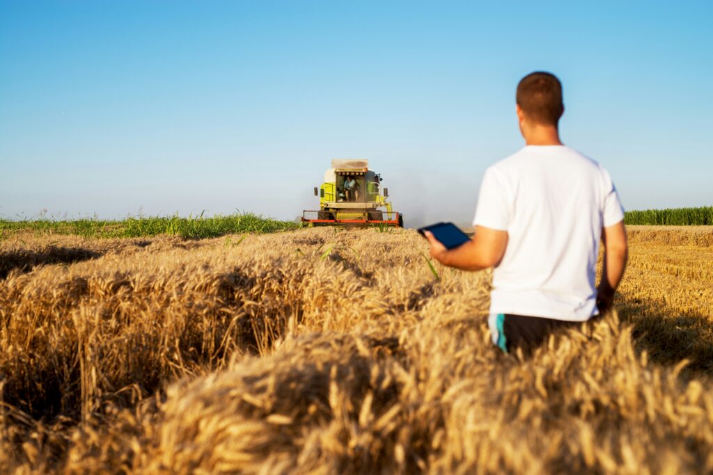 A farmer holding a tablet observes a combine harvester in a wheat field, illustrating agricultural equipment and machinery safety.