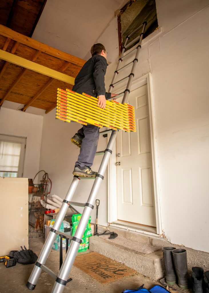 A worker climbs a telescoping ladder inside a garage while carrying safety equipment, representing defective ladder or product injury risk.