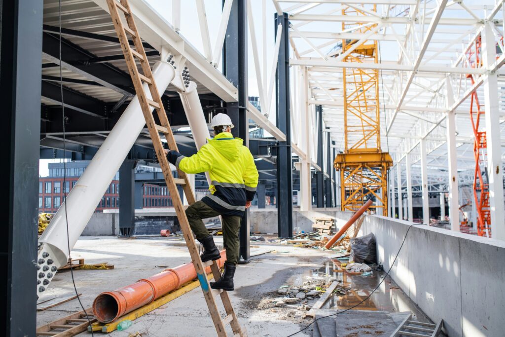 Construction worker in safety gear climbing a ladder on a busy commercial construction site with cranes and steel beams in the background.