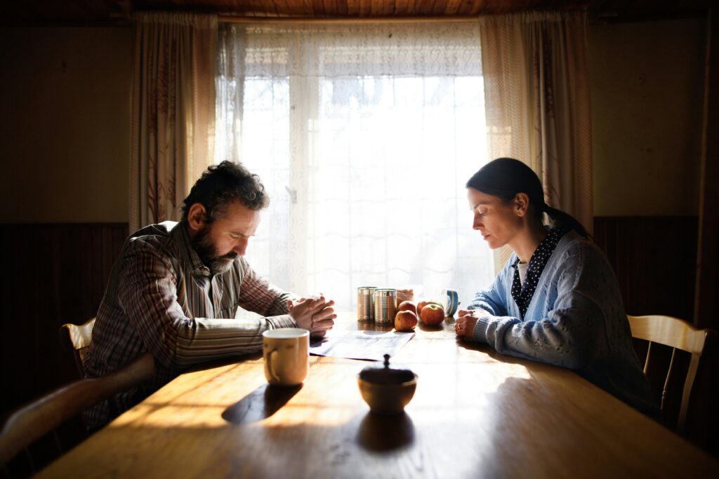 Grieving couple sitting at a kitchen table with heads bowed in sorrow, symbolizing loss and emotional pain after a loved one’s death.