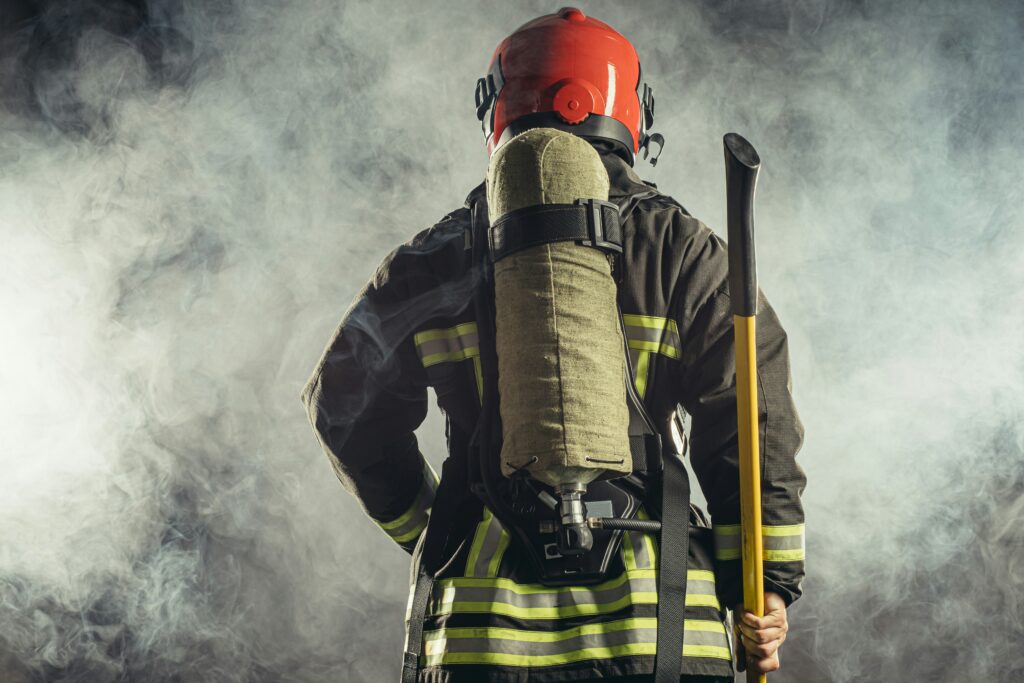Back view of a firefighter in helmet and turnout gear, wearing an air tank and holding an axe, standing in smoky haze.