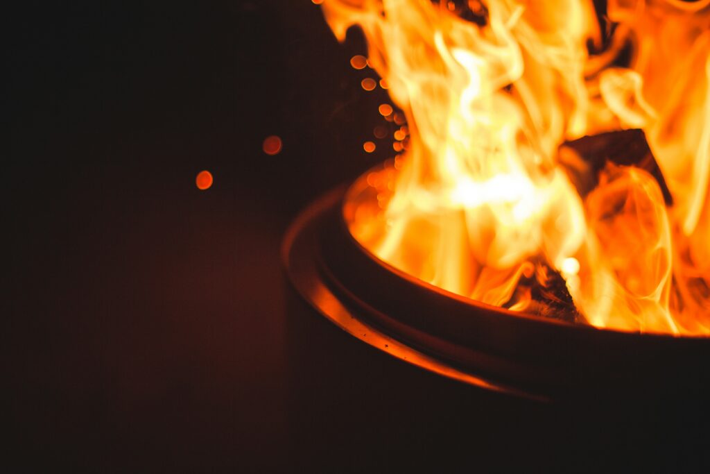Close-up of bright orange flames and sparks rising from a metal fire pit in the dark.