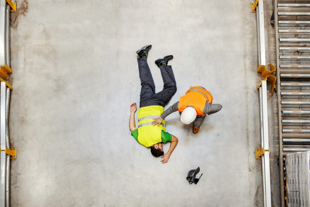A worker wearing a safety vest lies injured on a warehouse floor while a colleague in an orange vest kneels beside him to provide help.