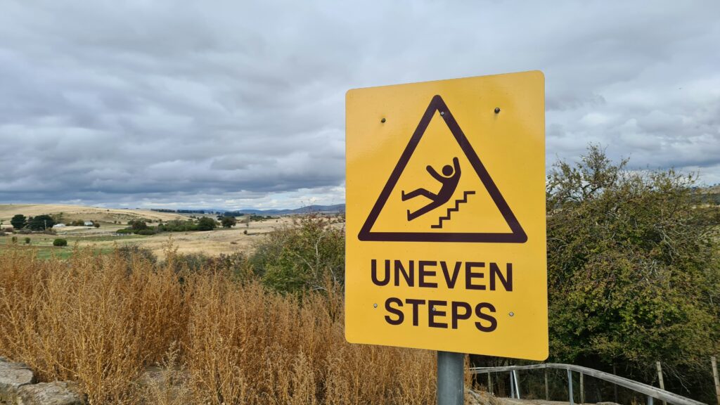 Yellow caution sign reading &ldquo;Uneven Steps&rdquo; beside an outdoor pathway under a cloudy sky, warning pedestrians of potential fall hazards.